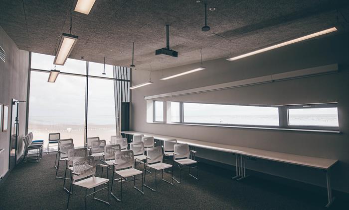 A wide angle view of a light function room. Chairs are spread in theatre style. A floor-to-ceiling window looking over a beach is in the background.